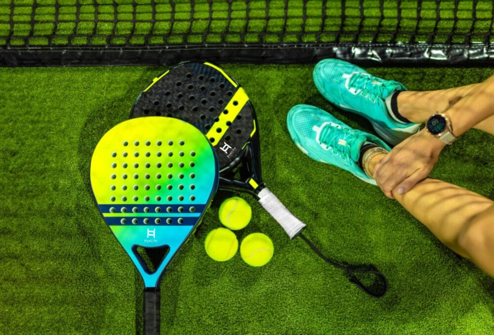 Custom branded paddle rackets and tennis balls on an indoor court beside a player wearing teal sneakers, showcasing custom sports equipment